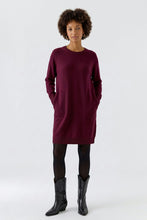 Load image into Gallery viewer, A woman modeling a long-sleeve crewneck knit dress in deep burgundy, paired with black tights and black leather ankle boots, against a plain light gray studio background.