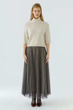 Load image into Gallery viewer, A woman stands against a light gray studio background wearing a cream short-sleeve mock-neck knit top and a long dark gray pleated tulle skirt with a subtle plaid pattern, paired with dark brown ankle boots.