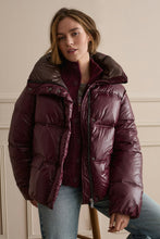 Load image into Gallery viewer, A woman wearing a shiny quilted puffer jacket in deep burgundy, styled over a matching knit top and light blue jeans, standing against a cream-colored paneled wall.