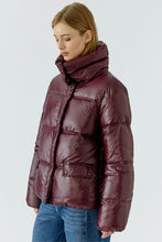 Load image into Gallery viewer, A woman wearing a shiny, quilted puffer jacket in a deep burgundy color, paired with blue jeans, photographed against a light gray studio background.