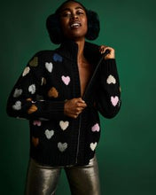Load image into Gallery viewer, A woman wearing a black knit zip-up cardigan with colorful heart motifs in pink, blue, yellow, and white, paired with metallic silver pants, set against a solid dark green background.