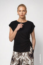 Load image into Gallery viewer, A woman modeling a black short-sleeve top with a gathered side detail, paired with patterned pants, against a light gray background.