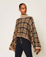 Load image into Gallery viewer, A woman modeling a chunky knit turtleneck poncho sweater with a brown, beige, and black pattern, paired with dark trousers, against a plain light background.