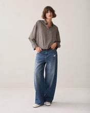 Load image into Gallery viewer, A model stands in a studio wearing a brown and beige vertically striped button-down shirt, wide-leg blue jeans, and white sneakers against a plain off-white background.