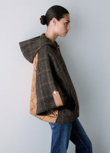 Load image into Gallery viewer, A woman in profile wearing a brown and black plaid hooded jacket with tan quilted side panels and blue jeans, photographed against a plain light gray studio background.