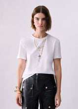 Load image into Gallery viewer, A woman modeling a white short-sleeve crewneck t-shirt, styled with layered gold necklaces, a gold cuff bracelet, and black leather cargo pants against a light gray studio background.