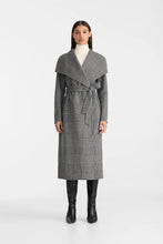 Load image into Gallery viewer, A model stands against a light gray studio background wearing a long gray plaid wrap coat with a wide collar and waist tie, over a white turtleneck and black knee-high boots.