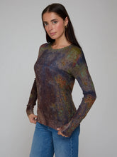 Load image into Gallery viewer, A woman modeling a long-sleeve crewneck knit top with an abstract, mottled multicolor pattern in shades of blue, green, brown, and gold, paired with blue jeans against a plain gray studio background.