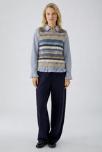 Load image into Gallery viewer, A model stands against a light gray studio background wearing a striped knit vest over a blue-and-white striped shirt, paired with dark navy trousers and black shoes.
