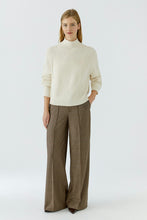 Load image into Gallery viewer, A woman modeling a cream mock-neck knit sweater and brown wide-leg trousers against a plain light gray studio background.
