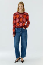 Load image into Gallery viewer, A woman modeling a long-sleeve crewneck sweater with an argyle pattern in red, orange, and burgundy, paired with blue jeans and dark brown pointed-toe heels, against a light gray studio background.
