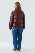 Load image into Gallery viewer, A woman viewed from the back wearing a shiny dark burgundy puffer jacket, blue jeans, and brown pointed-toe heels against a light gray studio background.
