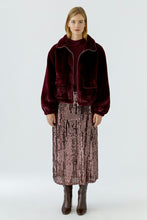 Load image into Gallery viewer, A model stands against a light gray studio background wearing a dark burgundy velvet zip-front jacket with a high collar, paired with a metallic rose-pink sequined midi skirt and dark brown ankle boots.
