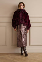 Load image into Gallery viewer, A model stands against a beige paneled wall wearing a deep burgundy velvet jacket over a matching burgundy turtleneck, paired with a metallic mauve sequined midi skirt and dark brown ankle boots.

