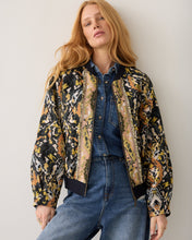 Load image into Gallery viewer, A woman wearing a black bomber jacket with a multicolor abstract print, styled over a blue denim shirt and jeans, posing against a plain light gray wall.
