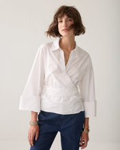 Load image into Gallery viewer, A woman wearing a white wrap-style blouse with three-quarter sleeves and navy pinstriped trousers, standing against a plain light gray studio background.
