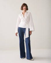 Load image into Gallery viewer, A model wearing a white long-sleeve wrap-style blouse with a tied waist and blue wide-leg jeans, standing against a plain light gray studio background.
