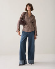 Load image into Gallery viewer, A model stands against a plain light beige studio background wearing a brown and beige vertically striped long-sleeve shirt with a tie-waist detail, blue wide-leg jeans with rolled cuffs, and carrying a dark brown shoulder bag.

