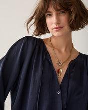 Load image into Gallery viewer, A close-up of a woman wearing a navy blue blouse and layered gold necklaces, including one with a silver ring and a small red and gold pendant, against a plain light background.
