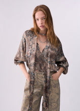 Load image into Gallery viewer, A woman modeling a loose, long-sleeve paisley blouse in beige, brown, and muted blue tones, paired with matching patterned pants, against a plain light gray studio background.
