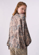 Load image into Gallery viewer, A model wearing a loose-fitting, long-sleeved paisley blouse with a beige, brown, and blue pattern, paired with matching patterned trousers, photographed against a light gray studio background.
