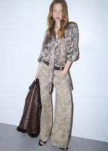 Load image into Gallery viewer, A model stands against a light gray studio wall wearing a matching beige and brown paisley print set consisting of a long-sleeve button-up shirt and wide-leg trousers, styled with a black belt and holding a dark brown leather jacket.
