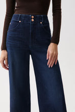 Load image into Gallery viewer, A close-up of a model wearing dark blue high-waisted wide-leg jeans with a double button closure and a brown ribbed long-sleeve top, set against a plain light gray background.
