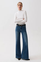 Load image into Gallery viewer, A woman modeling a white long-sleeve mock neck top and blue wide-leg jeans with a black belt, standing against a plain light gray studio background.
