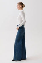 Load image into Gallery viewer, A woman in profile wearing a fitted white long-sleeve top and high-waisted, wide-leg blue denim jeans against a plain light gray studio background.
