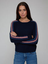 Load image into Gallery viewer, A woman modeling a navy blue knit sweater with red and white striped sleeve panels, paired with blue jeans, against a plain light gray studio background.
