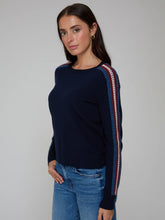 Load image into Gallery viewer, A woman wearing a navy blue knit sweater with red and white striped detailing on the sleeves, paired with blue jeans, photographed against a neutral gray studio background.
