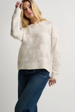 Load image into Gallery viewer, A woman wearing a cream-colored, long-sleeve knit sweater with an intricate openwork heart pattern, paired with dark blue jeans, against a plain light gray studio background.
