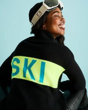 Load image into Gallery viewer, A woman wearing a black knit sweater with a bright neon yellow-green horizontal stripe across the back, printed with the word 'SKI' in teal. She is also wearing ski goggles on her head and a black beanie, set against a solid light blue background.
