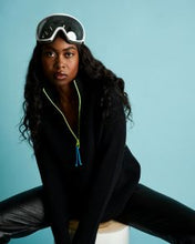 Load image into Gallery viewer, A woman wearing a black knit zip-up hoodie with a bright neon green drawstring and blue tassel, black leather pants, and white ski goggles resting on her head, posed against a solid light blue background.
