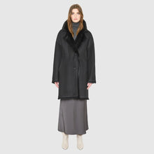 Load image into Gallery viewer, A woman modeling a black mid-length coat with a dark fur-trimmed collar, worn over a gray turtleneck and a long gray skirt, against a light gray studio background.
