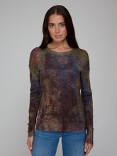 Load image into Gallery viewer, A woman modeling a long-sleeve crewneck knit top with a mottled, abstract multicolor pattern in shades of purple, brown, green, and blue, paired with blue jeans against a solid gray studio background.
