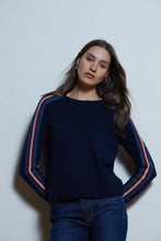 Load image into Gallery viewer, A woman wearing a navy blue knit sweater with red and white striped sleeve details, paired with dark blue jeans, standing against a plain light gray wall.
